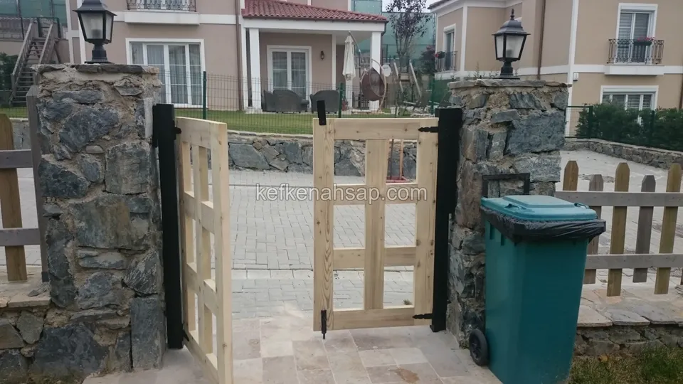 Taş sütunlar arasında, bir kanadı açılmış bölmeli açık renk ahşap çift kanatlı kapı; arkada taş duvar, ev ve çöp konteyneri