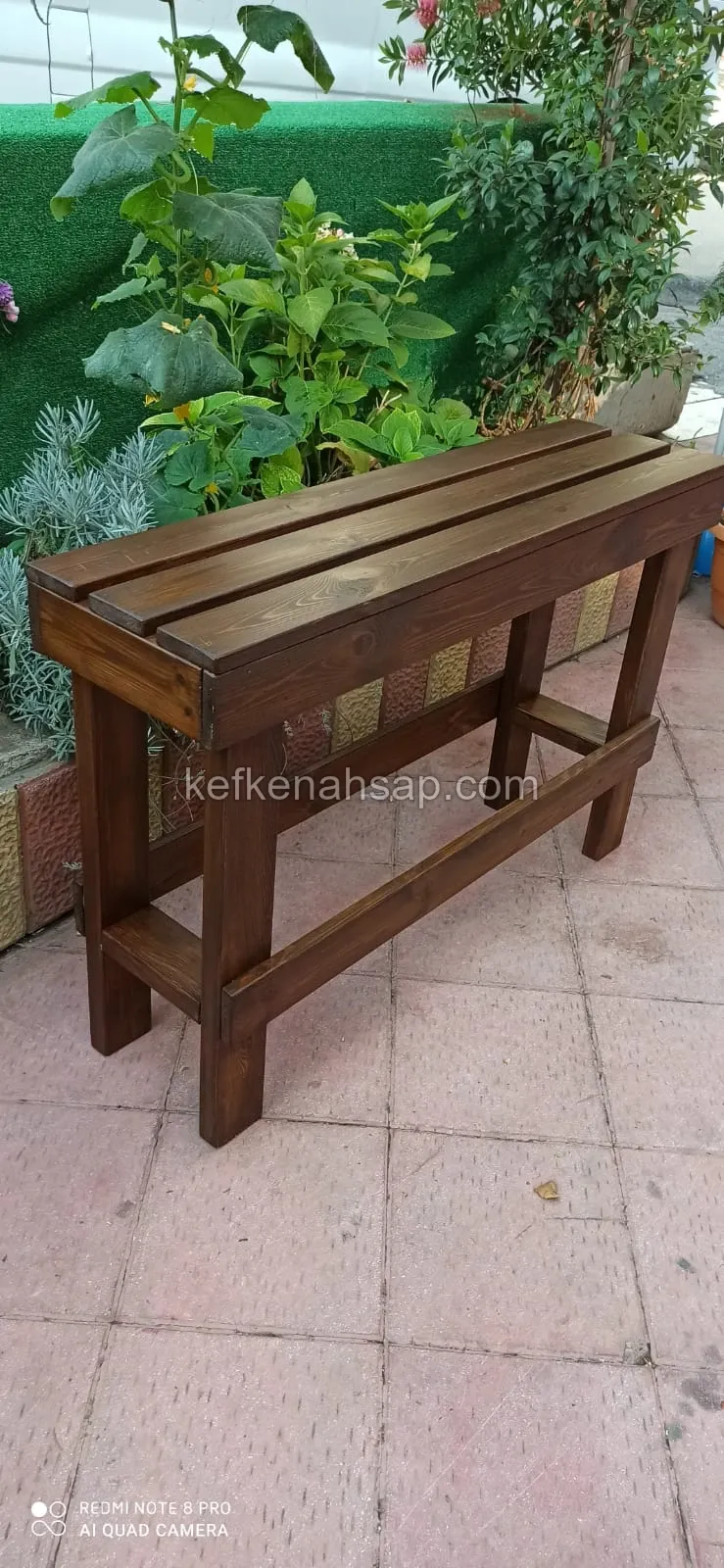 Dark brown stained wooden bench with three seat slats and lower rails, standing on tiled ground in front of plants