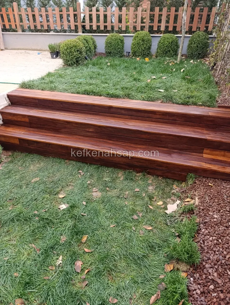Two-tier dark brown wooden garden steps up to a lawn with trimmed boxwoods, a wooden fence, grass and red gravel around