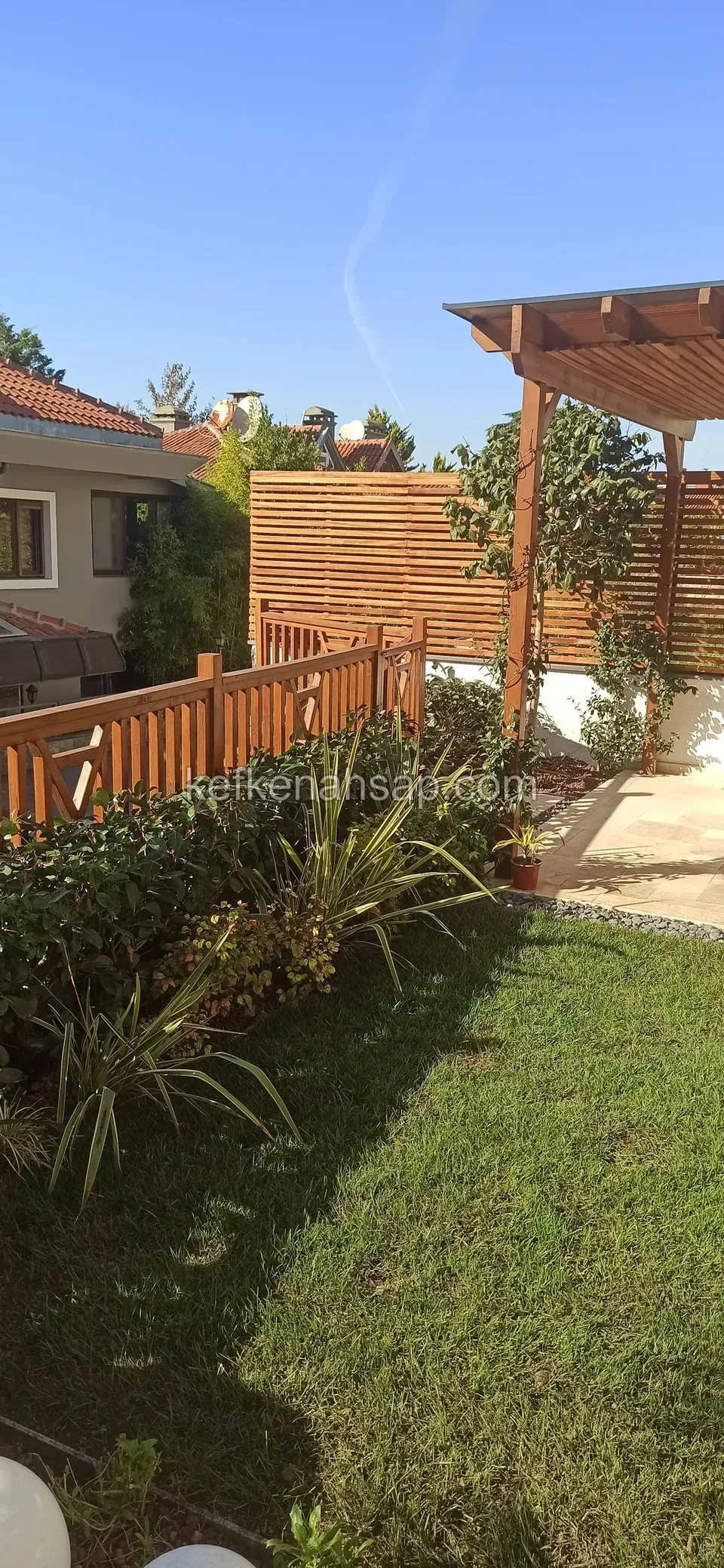 Garden lawn with a wooden railing fence, horizontal slat screen and a pergola over a stone-paved terrace
