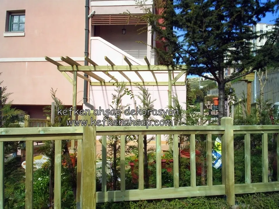 Pembe bir binanın önünde, dikmeler arasında dik çıtalı açık renk ahşap bahçe çiti ve arkasında ahşap pergola