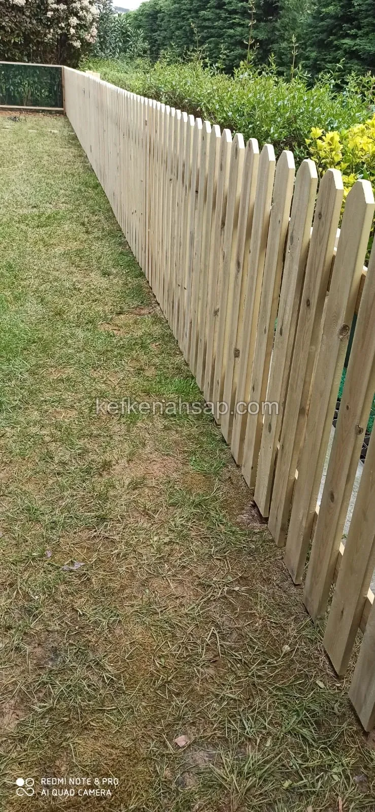 Çim bahçenin kenarında uzanan, uçları yuvarlatılmış dikey tahtalardan yapılmış açık renkli ahşap çit
