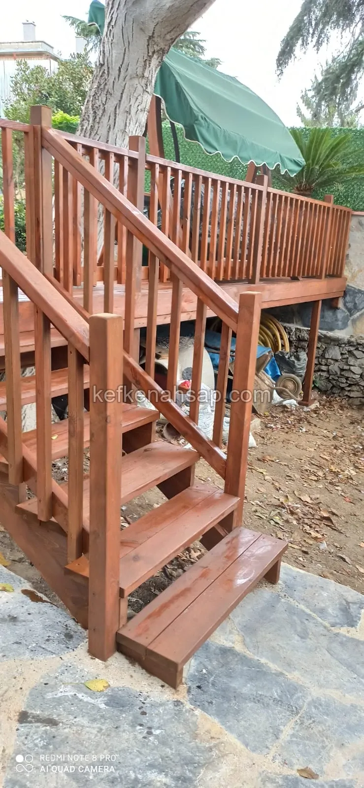 Bahçede ağacın çevresine kurulmuş, kızıl kahve cilalı ahşap deck, korkuluk ve taş zemine inen basamaklar