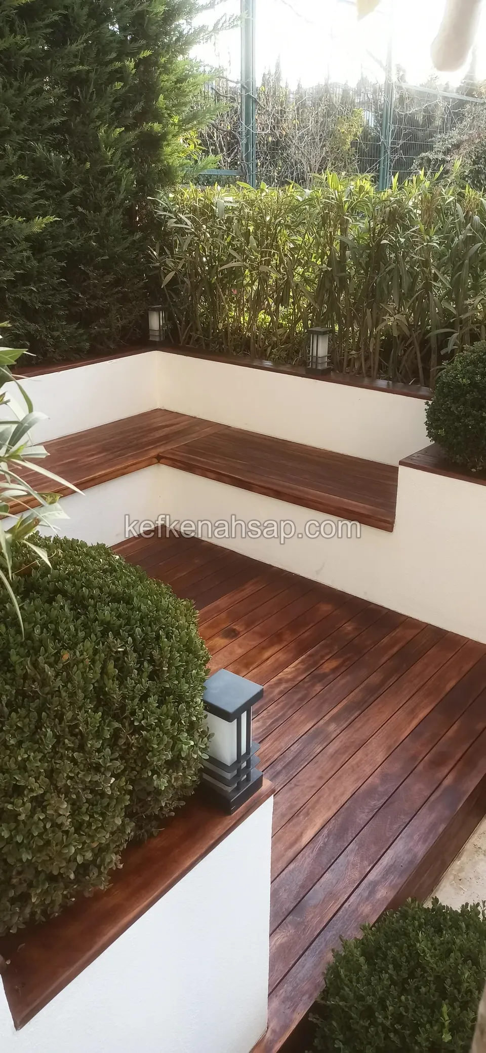 Dunkelrötliches Holzdeck in einer Gartenecke mit weißen Wänden, Sitzbank mit demselben Holz verkleidet, Gartenleuchten