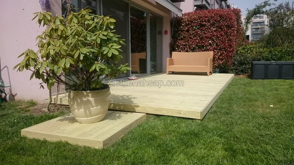 Çim bahçede veranda önüne kurulmuş açık renkli ahşap deck platformu ve saksılık basamağı.