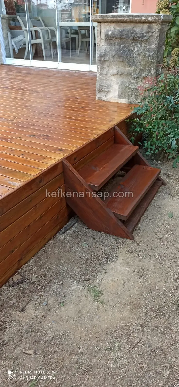Cam sürgülü kapının önünde kahverengi ahşap deck, yan cephesi ve toprak zemine inen iki basamaklı ahşap merdiven