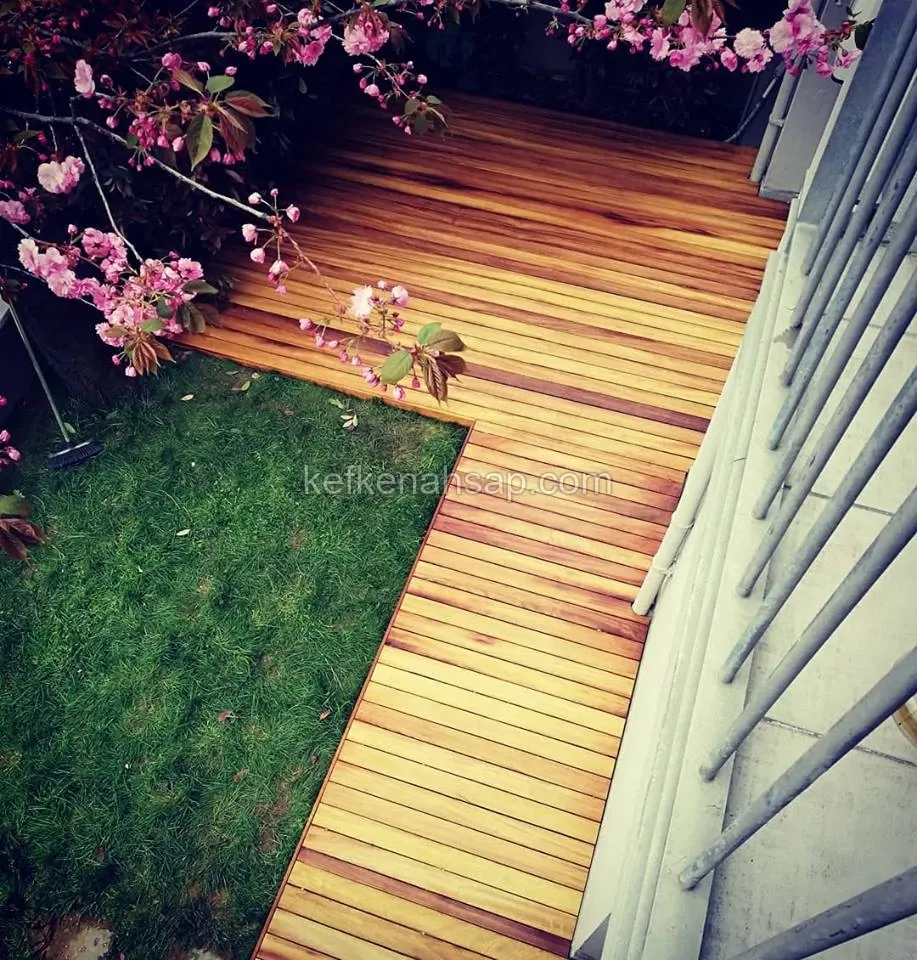 Çimenlik bahçede L şeklinde uzanan bal rengi ahşap deck, solda çiçek açmış pembe ağaç dalları, sağda metal korkuluk
