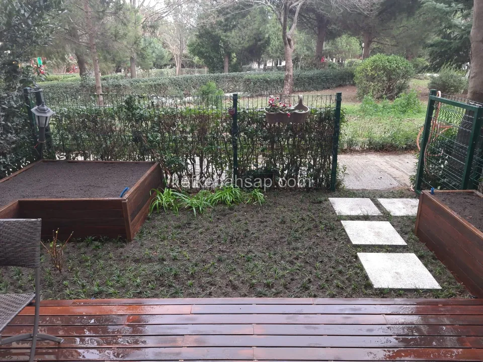 Bahçede ıslak kırmızımsı ahşap deck, yanında ahşap yükseltilmiş bitki yatakları, taş basamak plakaları ve yeşil telli çit