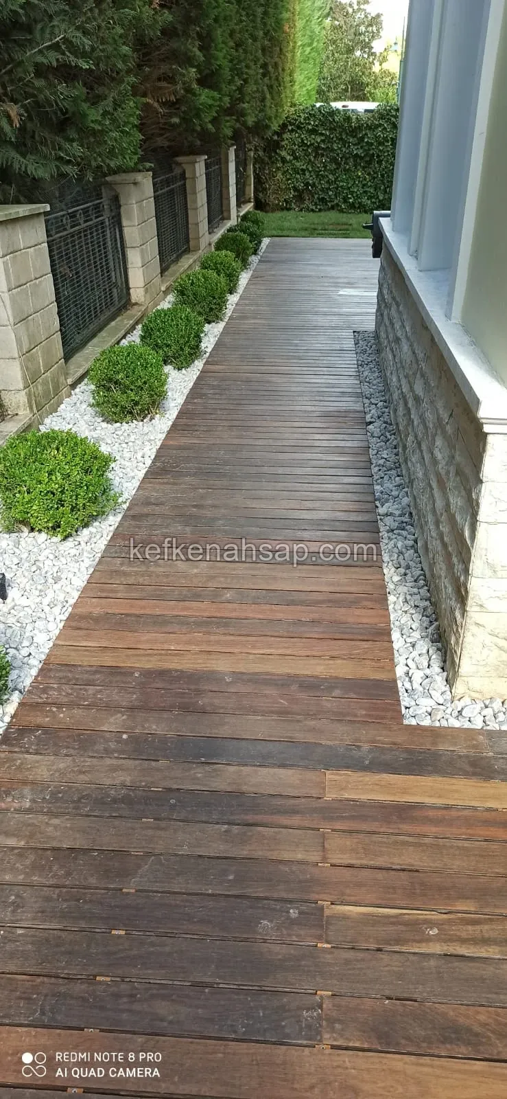 Bina kenarındaki bahçe boyunca uzanan ahşap deck yürüyüş yolu ve yanındaki beyaz çakıl taşları.