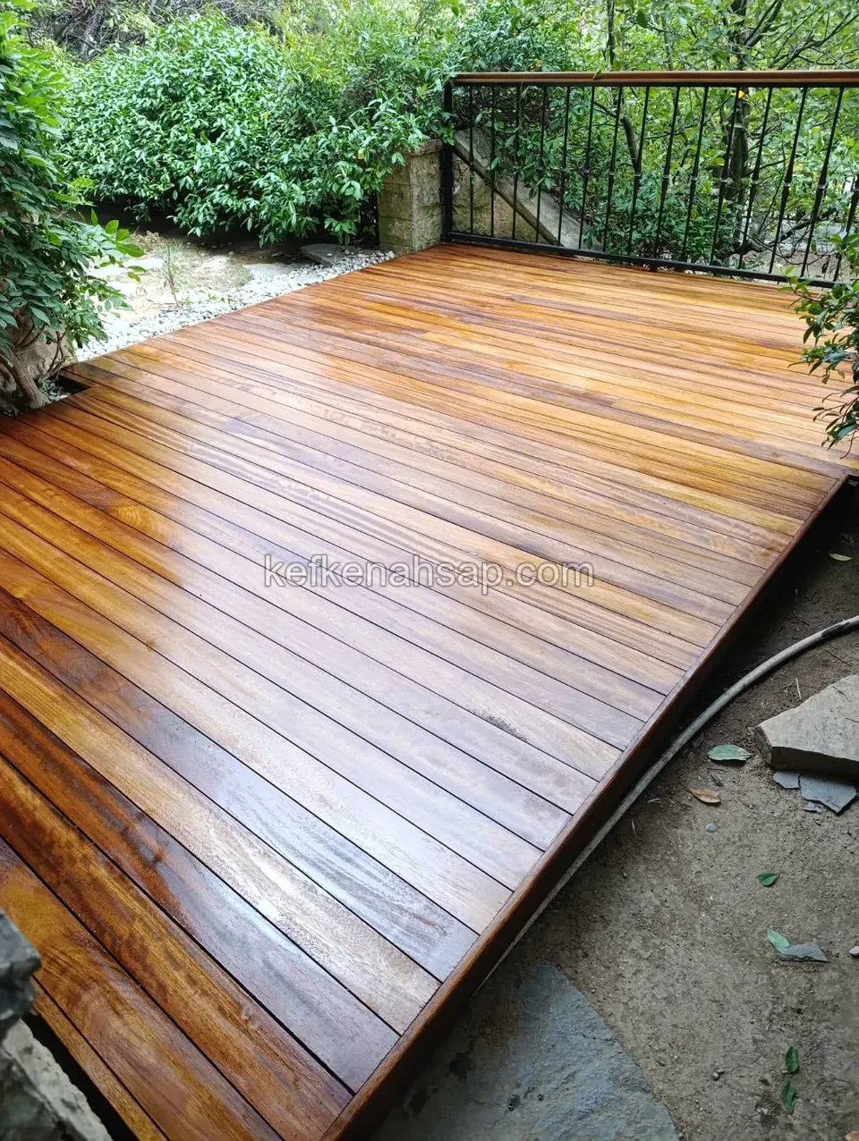 Yeşilliklerle çevrili bahçede, bal rengi ahşap tahtalarla döşenmiş deck zemin ve arkasında metal korkuluk