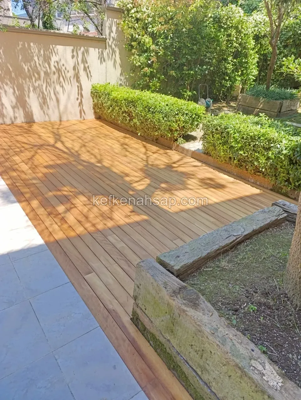 Narrow-board wooden deck in a garden between a wall and hedges, lit by sun and tree shadows