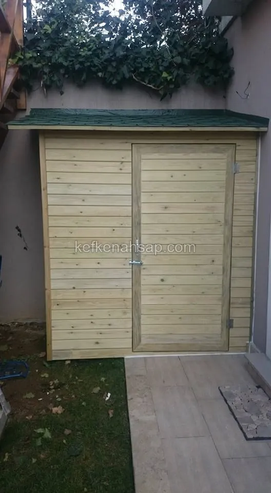Bahçede duvara yaslanmış, yeşil shingle çatılı, yatay kaplamalı ve tek kapılı açık renk ahşap depo