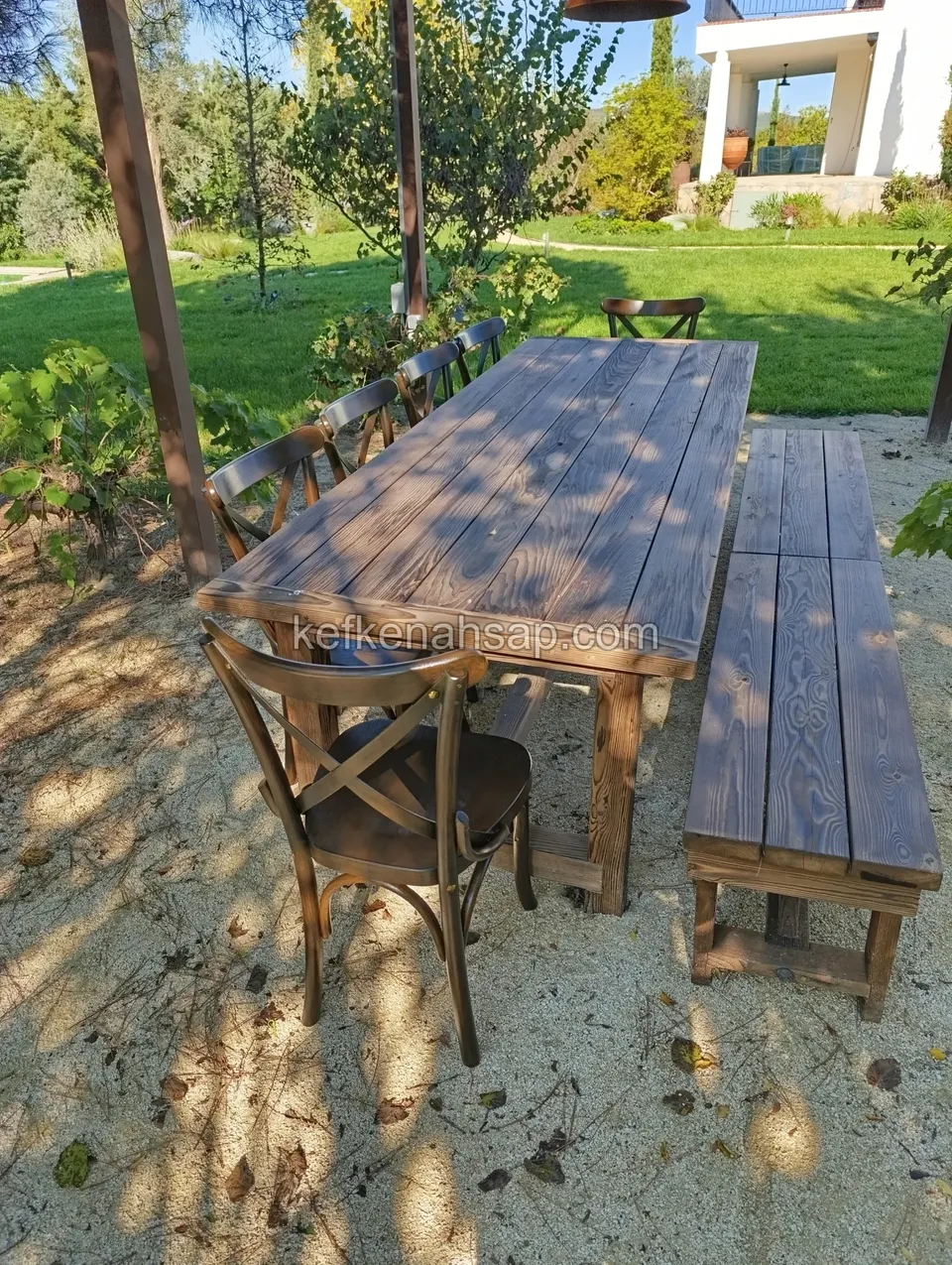 Long plank wooden table on a gravel garden floor with a bench and cross-back chairs beside it