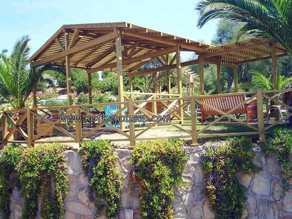 Palmiye ve çiçeklerin arasında, çapraz çıtalı korkuluğu olan, çatısı ahşap çıtalarla kaplı iki bölümlü ahşap pergola