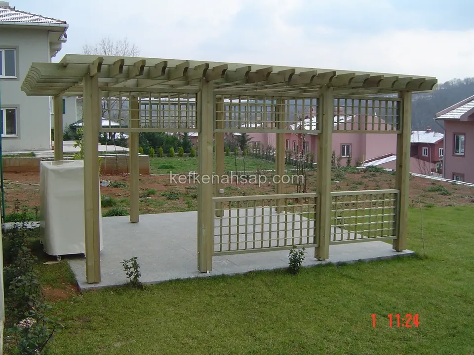 Taş kaplı zemin üzerinde kafes panelli, üstü kirişli açık renkli ahşap pergola; arkada villalar ve çim alan