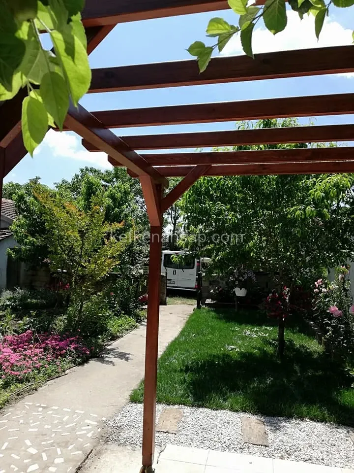 Bahçe yolunun üzerinde kahverengi boyalı ahşap pergola, kirişlerin ardında çim alan, çiçek tarhı ve ağaçlar