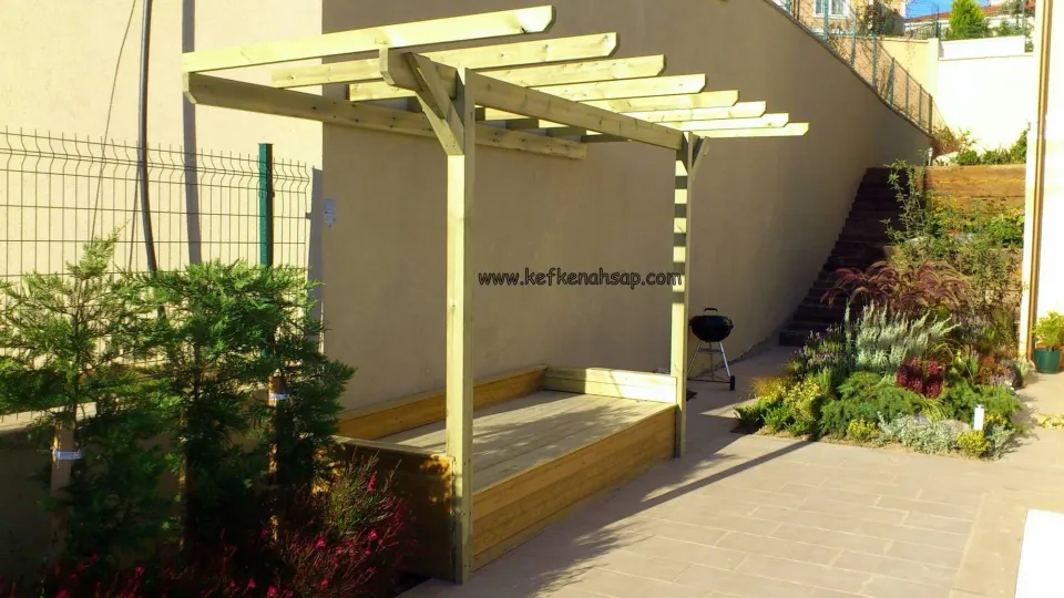 Bahçe duvarının önünde açık renk ahşap pergola, altında ahşap kaplı oturma platformu, yanında mangal ve bitkiler