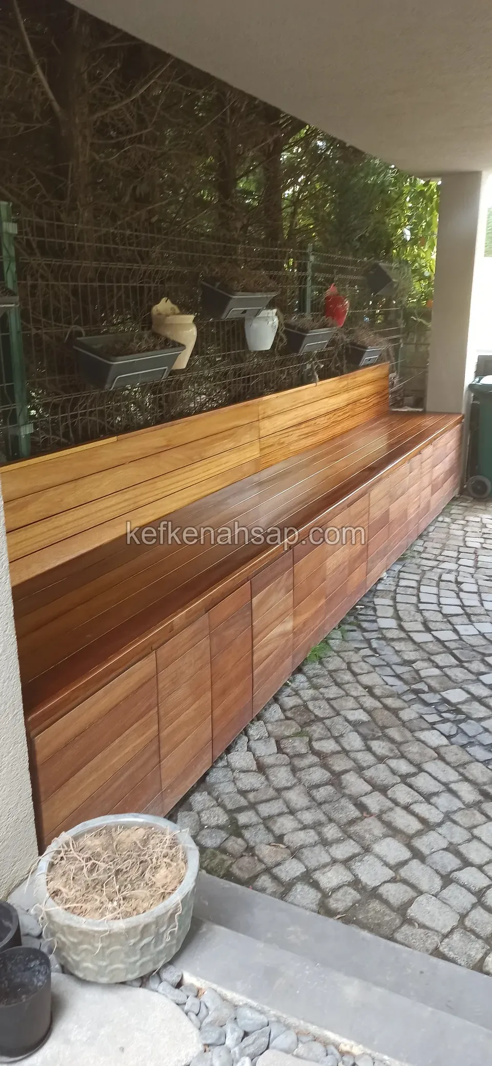 Long wooden bench with backrest and cabinet front beside a cobblestone garden path, planters on the fence behind