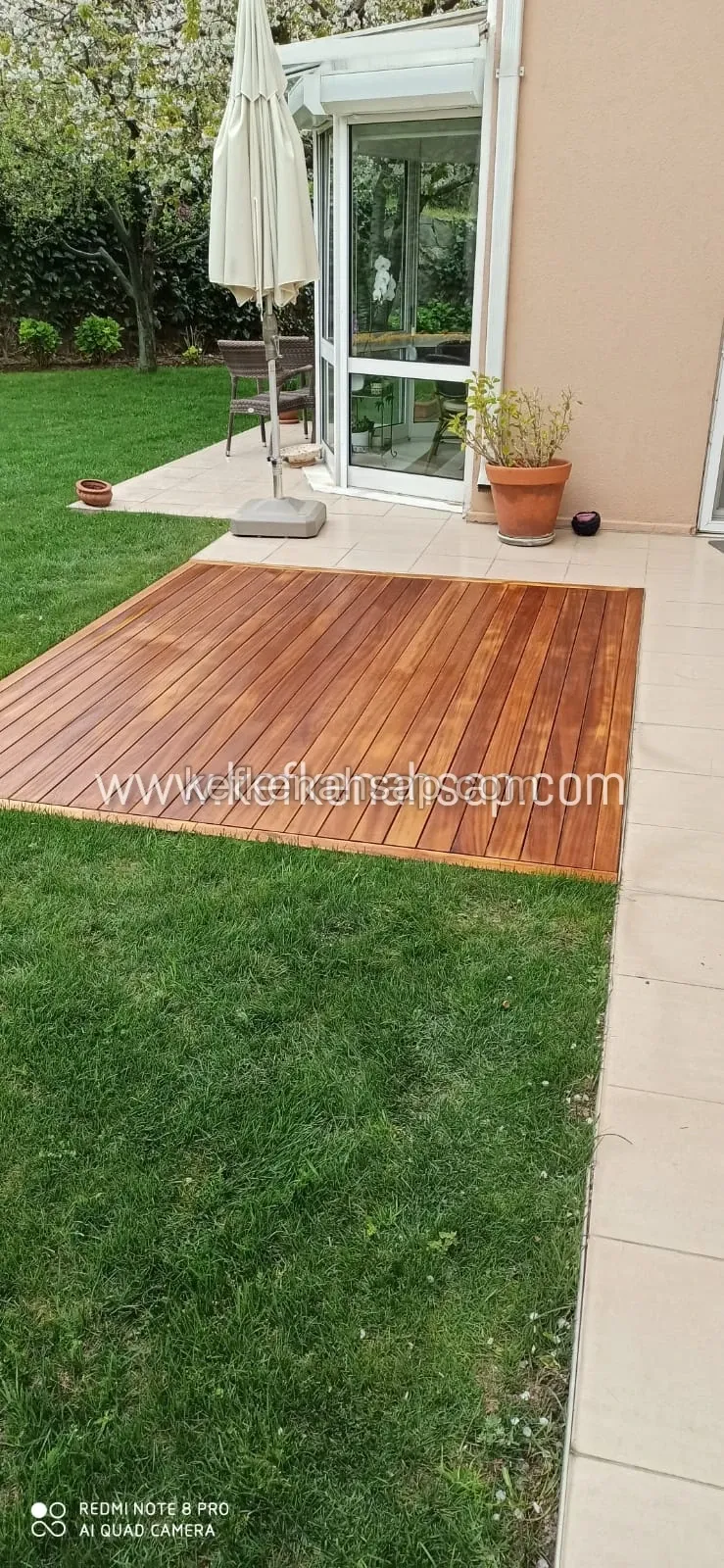 Bahçede çimenlerin arasına döşenmiş, bal rengi ahşap çıtalı kare deck; arkada cam kapılı teras ve şemsiye