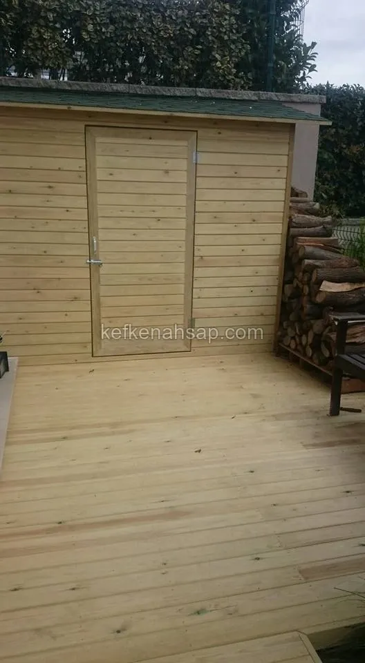 Yeşil çatılı ahşap bahçe deposu, yatay kaplamalı kapısı ve önünde deck zemin; yanda istiflenmiş odun kütükleri
