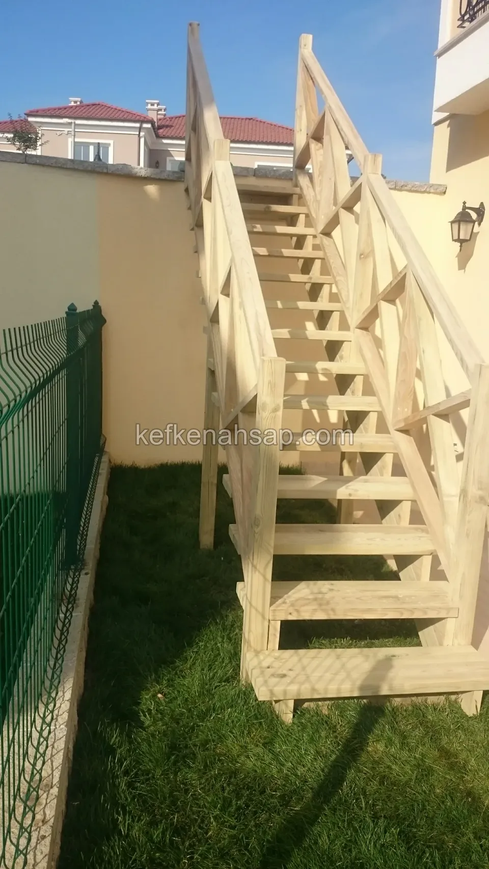 Holztreppe mit Kreuzstreben-Geländern entlang einer beigen Gartenmauer, links grüner Maschendraht, an der Wand eine Lampe