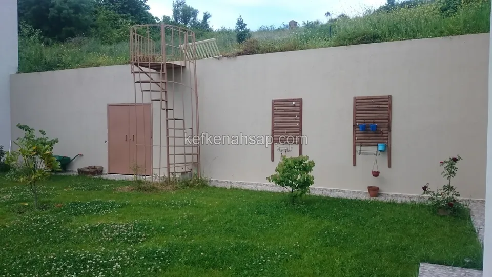 Krem bahçe duvarında açık kahve ahşap çift kanatlı kapı, iki küçük jaluzi panel ve yanındaki spiral merdiven, önde çimenlik