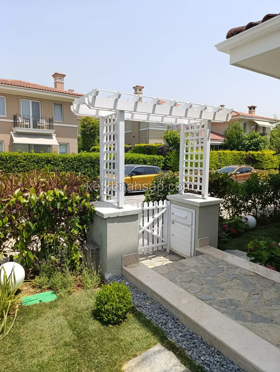 Taş kolonlar üzerinde beyaz kafes yan panelli ahşap pergola, yanında beyaz bahçe kapısı ve taş döşeli giriş yolu