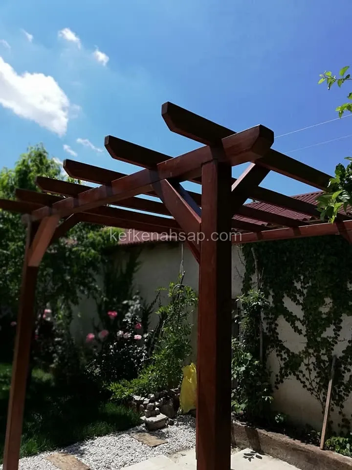 Bahçede kızıl kahve vernikli ahşap pergola, çapraz destekler ve üstte sıralı kirişler, arkada taş duvar ve sarmaşık