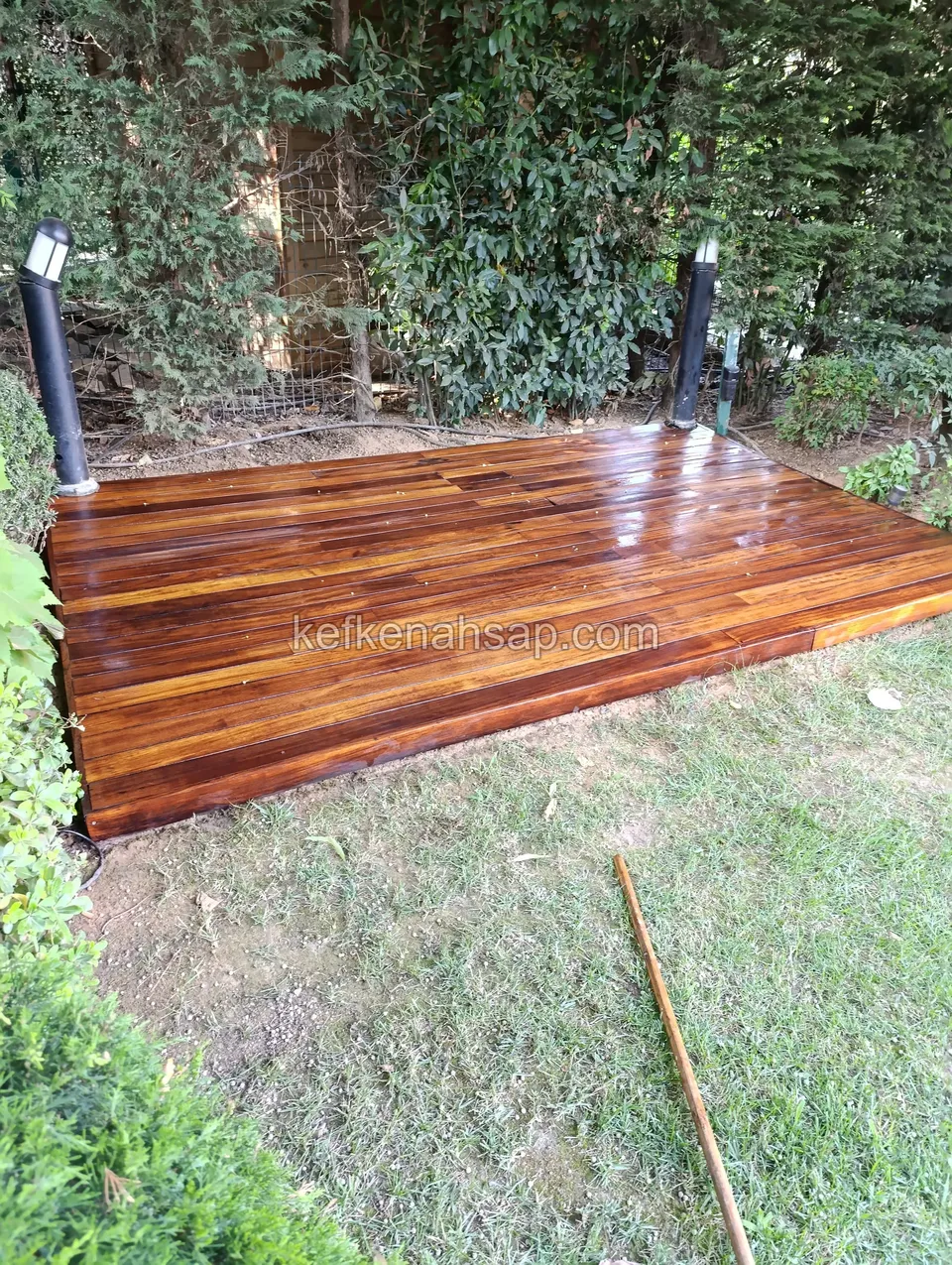 Small glossy dark red-brown deck platform set on the lawn in front of a hedge between two garden lamps