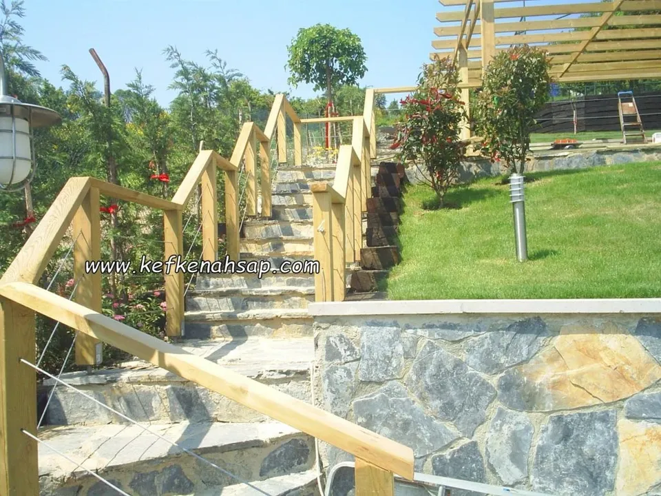 Eğimli bahçede taş basamaklı merdivenin yanında çelik halat dolgulu, açık renk ahşap korkuluk ve arkada ahşap pergola