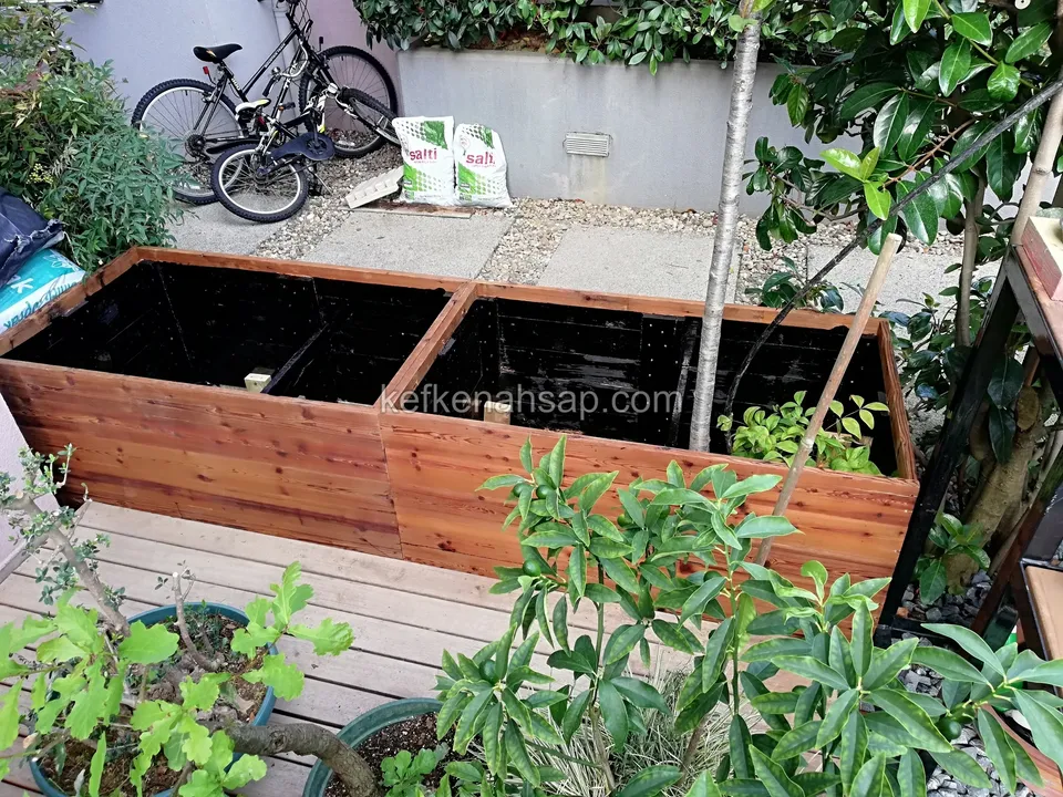 Bahçe terasında iki bölmeli ahşap çiçeklik; arkada bisikletler, saksılar ve beton duvar