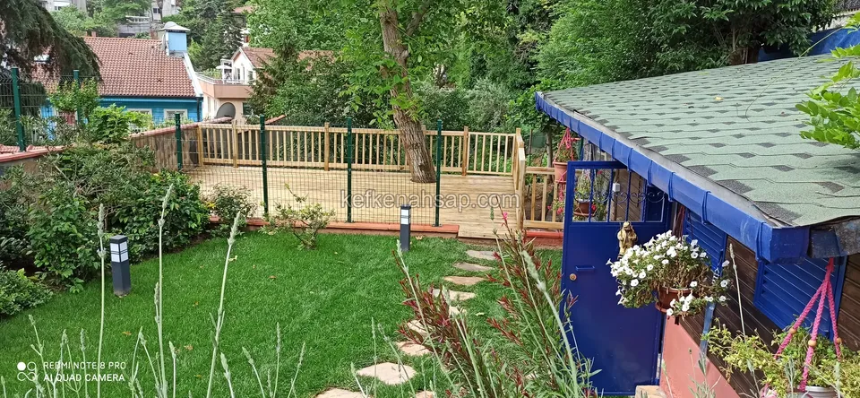 Çimenli bahçenin ucunda dikey çıtalı ahşap korkulukla çevrili ahşap deck ve yanında mavi kapılı bahçe evi