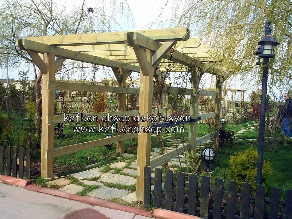 Taş döşeli bahçe yolunun üzerinde, yeşilimsi emprenyeli ahşaptan yapılmış, yan korkuluklu ve kirişli pergola