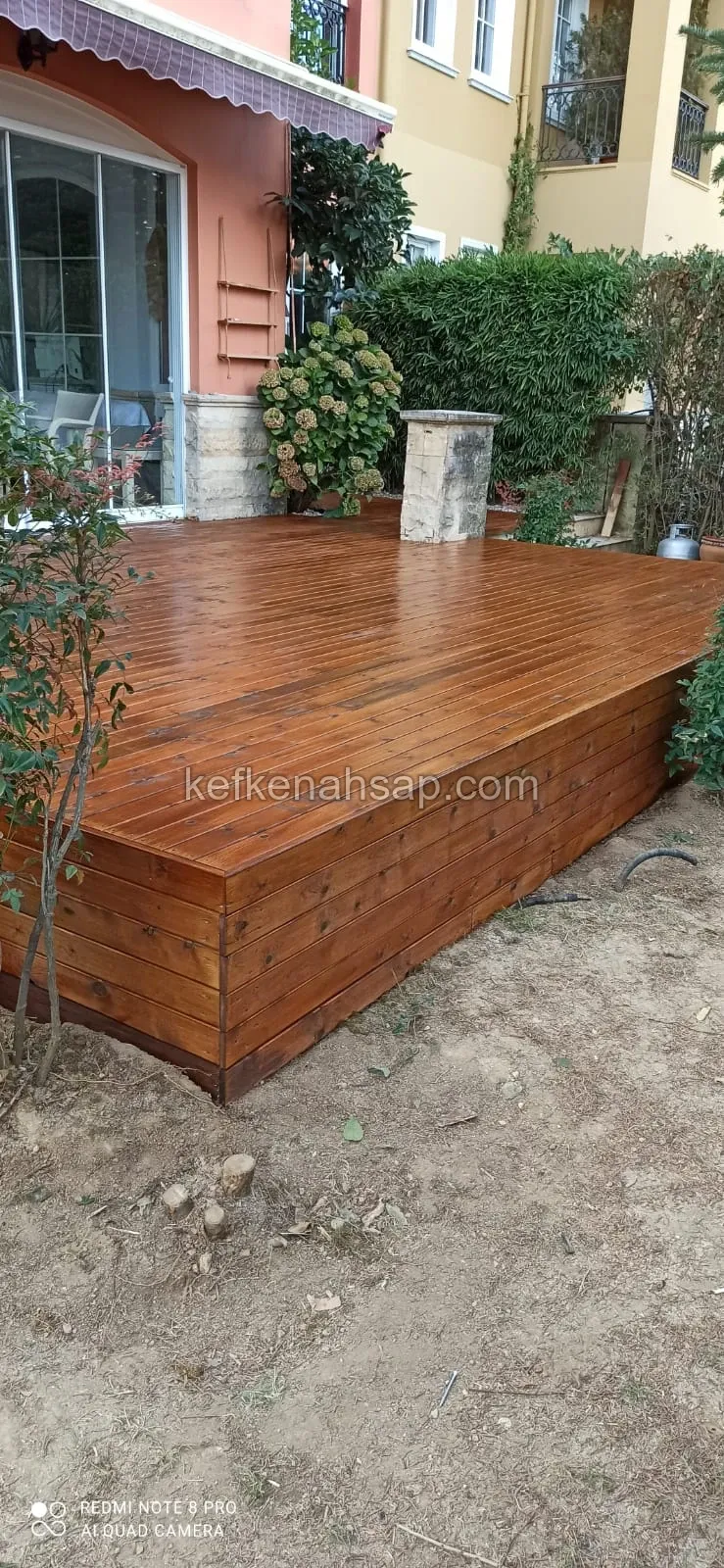 Pembe cepheli bir binanın bahçesinde, cam kapının önünde yan yüzeyleri de ahşap kaplı, yükseltilmiş bal rengi deck