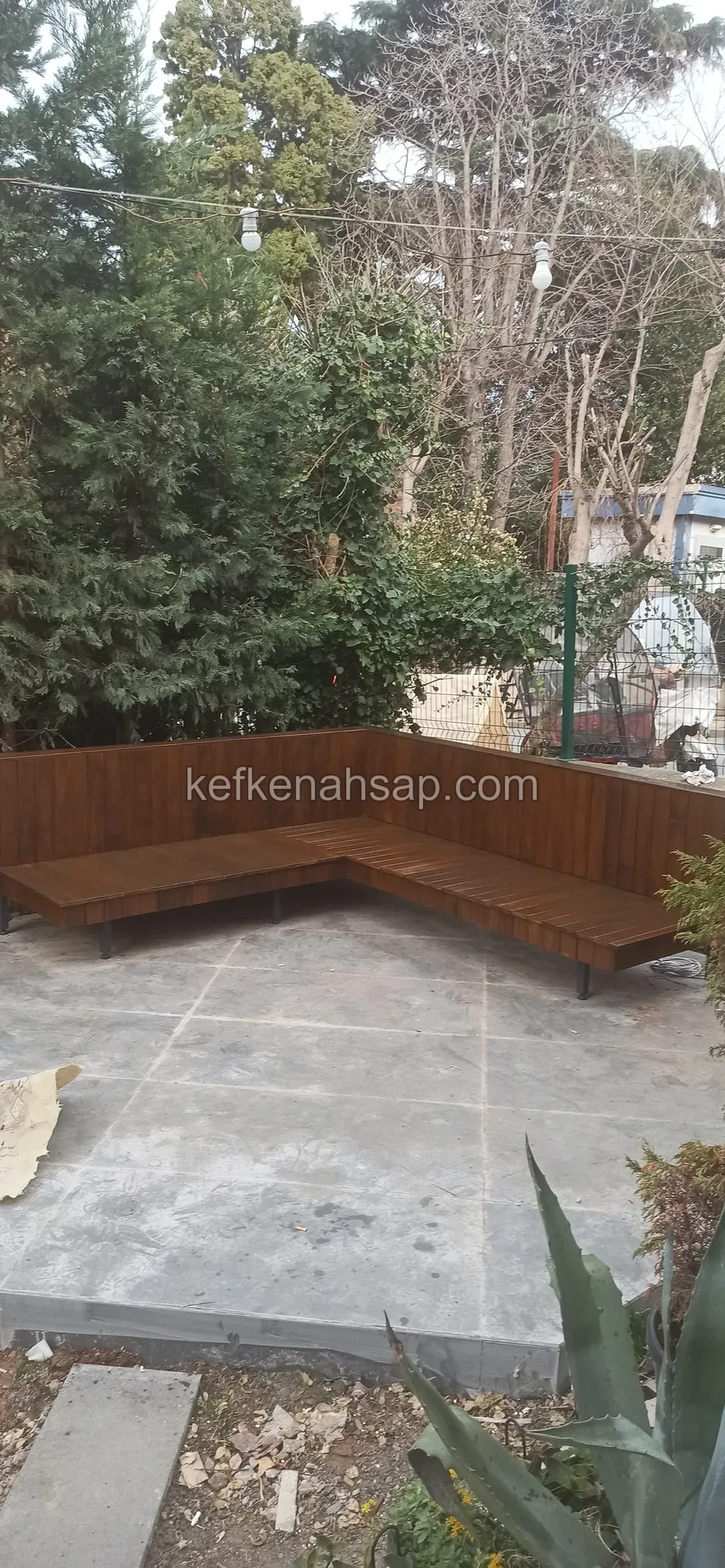 Aus der Ferne sieht man eine dunkle L-förmige Holzbank entlang eines Zauns in einem Garten mit Betonboden
