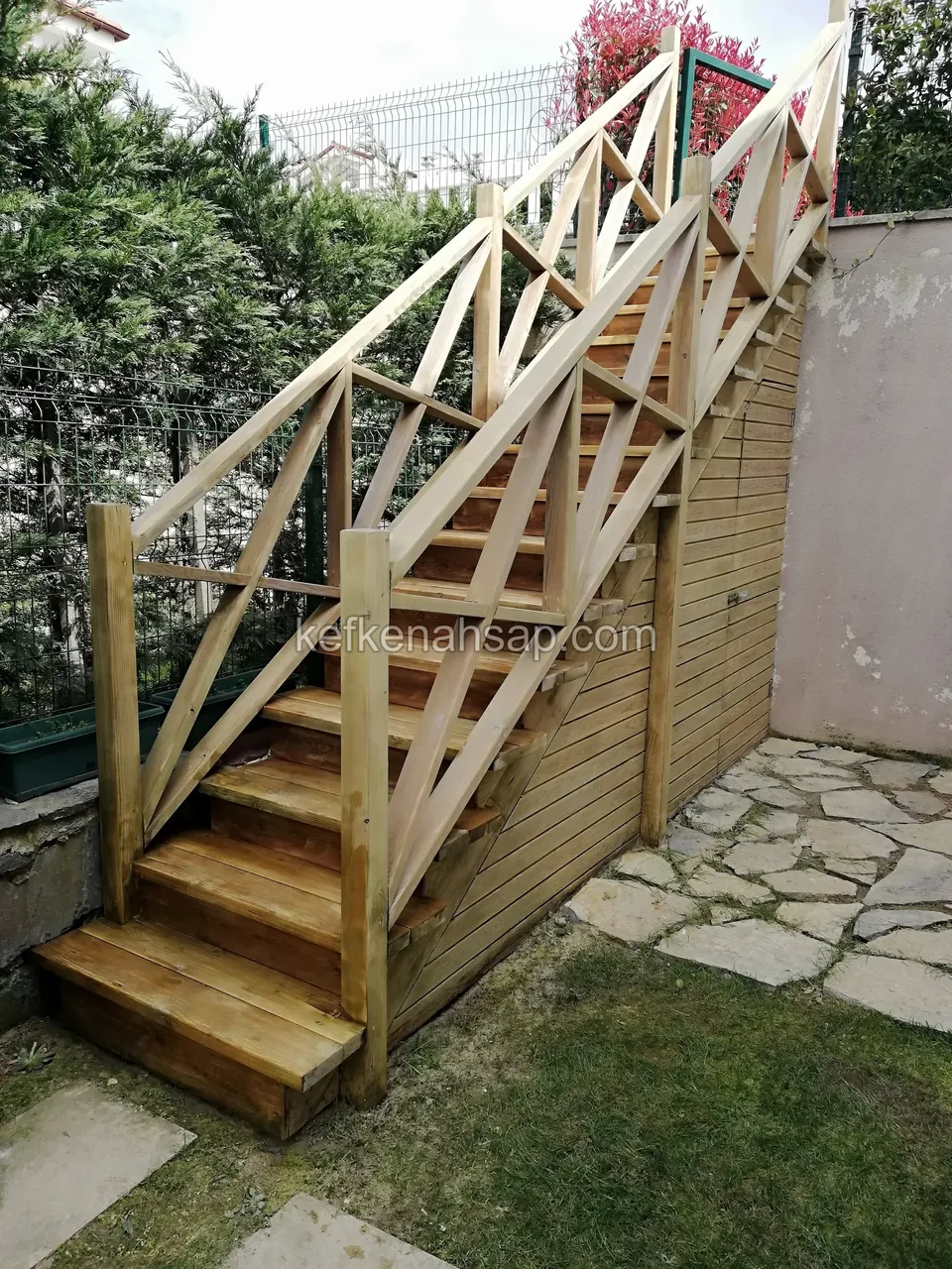 Gartentreppe mit Diagonalgeländer auf Rasen und Steinbelag, darunter ein holzverkleideter Schuppen mit Tür
