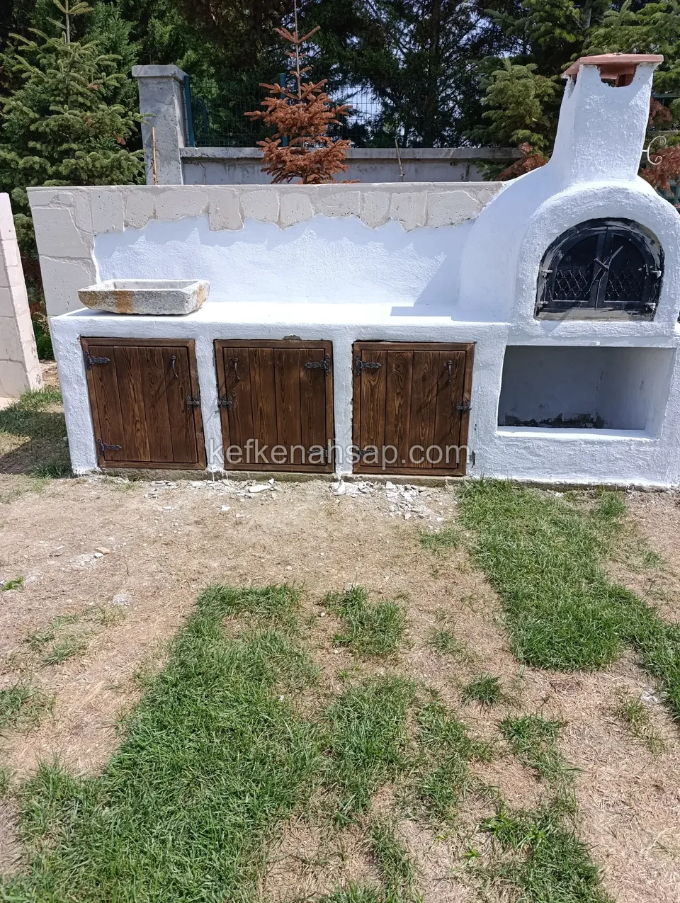 Bahçede, beyaz boyalı bloktan örülmüş ocaklı fırın ve altında üç ahşap kapaklı dolap