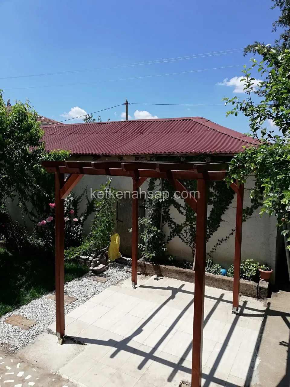 Taş zeminli bahçede koyu vernikli ahşap pergola, arkada kiremit rengi çatılı bina ve sarmaşıklı duvar