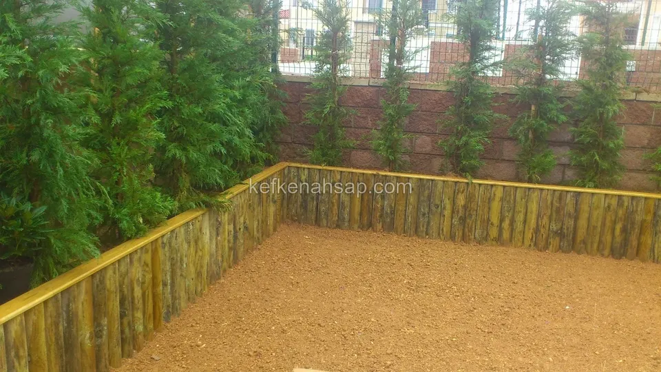 Low border made of log slats in a garden, with a sandy area in the middle and cypress trees behind