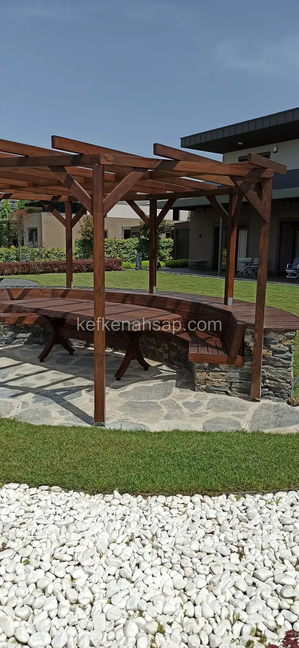 Çimlik bahçede yarım daire taş duvarlı oturma alanının üstünde koyu vernikli pergola, arkada modern bir ev