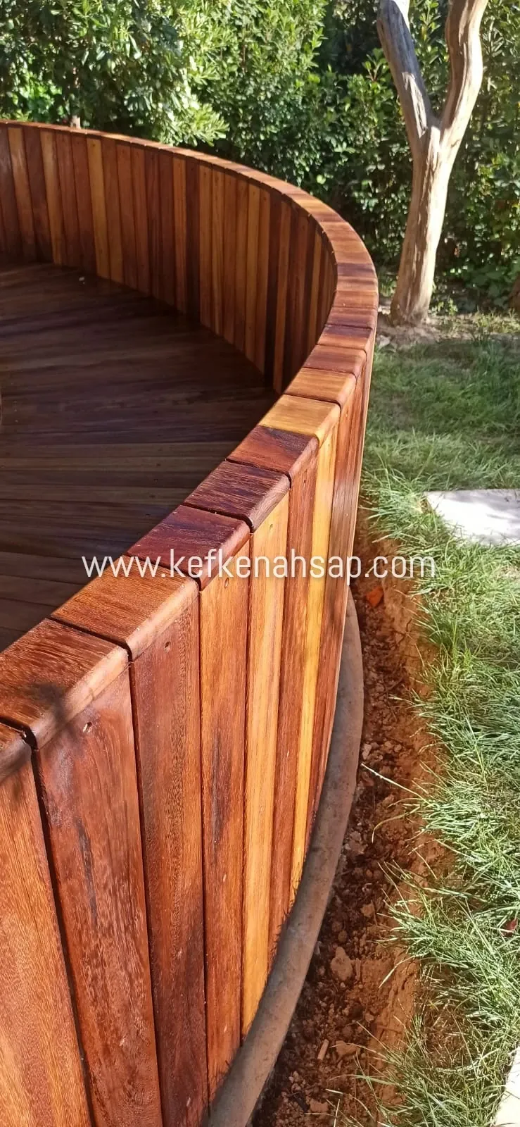 Bahçede yuvarlak bir ahşap platformun dikey çıtalarla kaplı kenarı, üstünde ahşap döşeme, yanında çimen ve ağaç