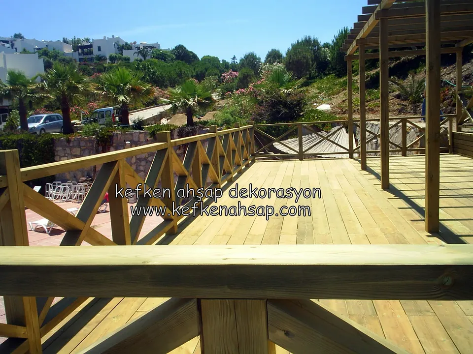 Palmiyeli bir bahçeye bakan geniş deck zemini, çapraz korkuluk, kalın tutamak kirişi ve pergola kolonları