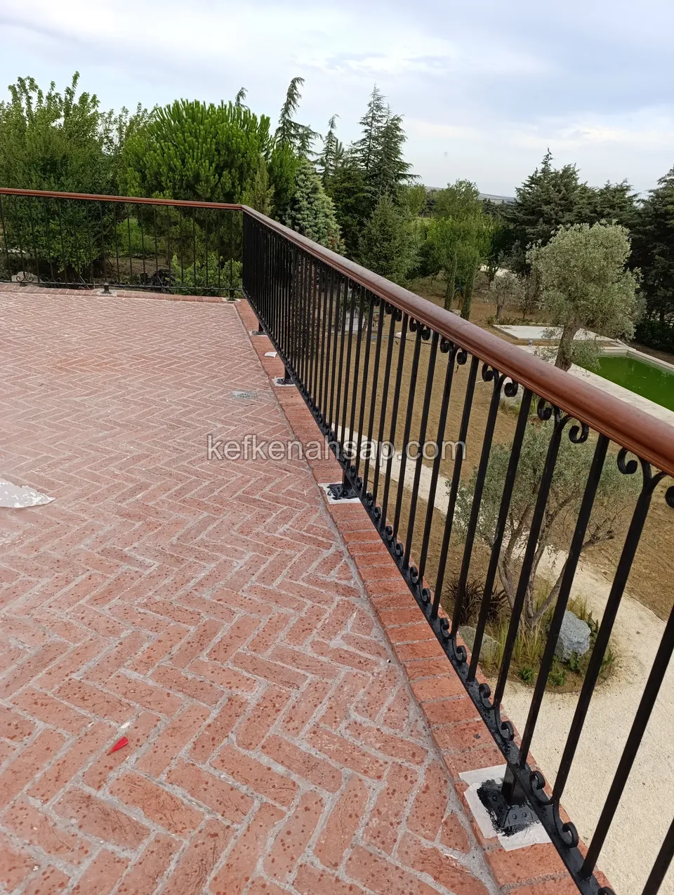 Black iron railing with curled decoration and a brown timber handrail along a terrace paved in red herringbone brick