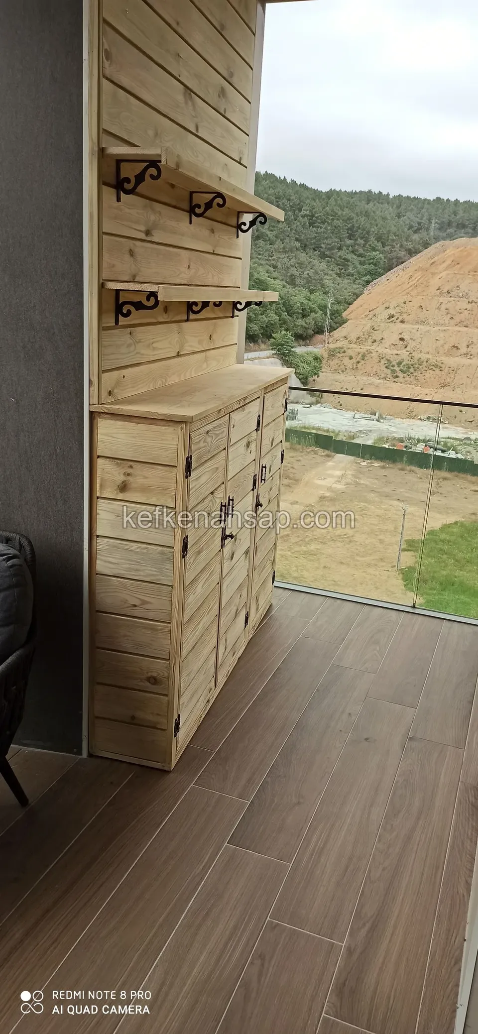 Zwei Regale auf schwarzen Konsolen an einer mit Brettern verkleideten Wand auf einem Balkon, darunter ein Schrank mit Türen