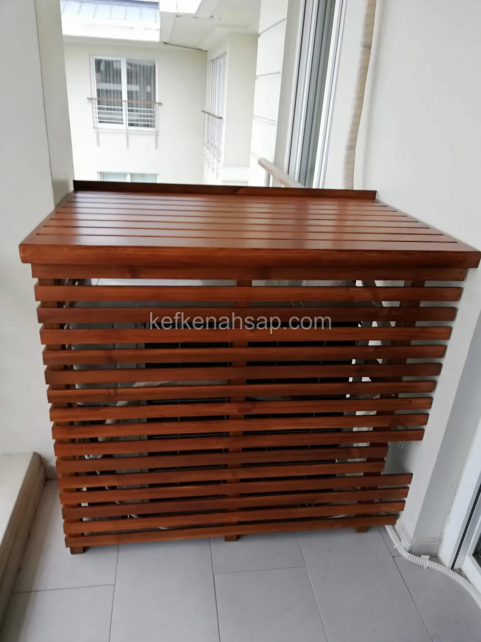 Red-brown timber air-conditioner housing with spaced horizontal slats on the sides and a boarded top in a balcony corner