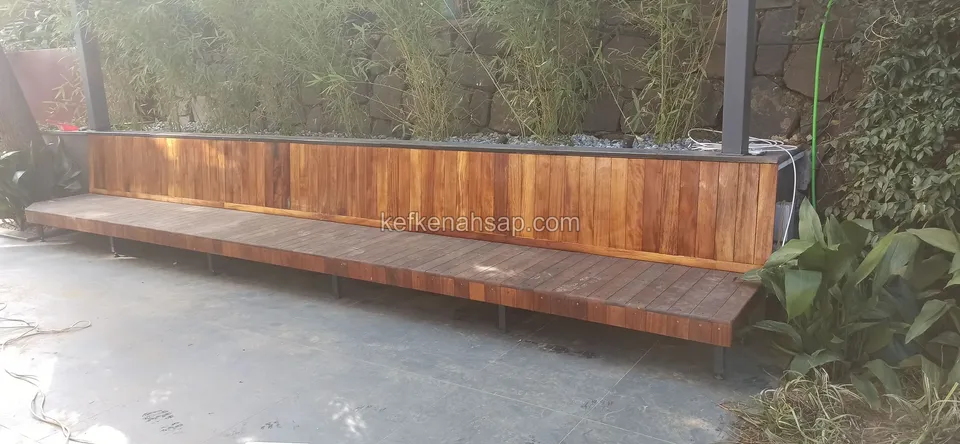 Beton zeminde, alçak bir bambu bahçesi ve taş duvarın önünde uzanan, dikey şeritli arkalığı olan çok uzun ahşap bank