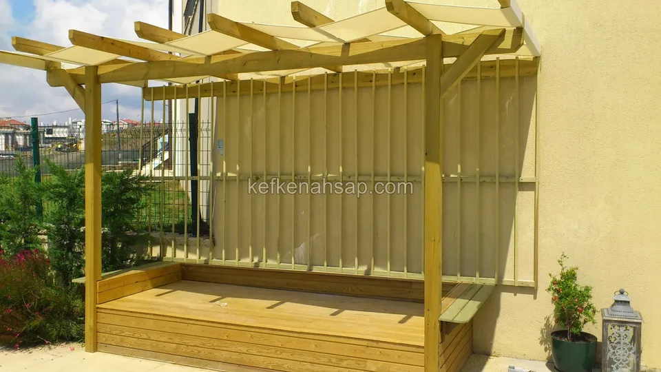 Sarı sıvalı duvar önünde, dikey çıtalı arka paneli ve ahşap oturma bankı olan pergola