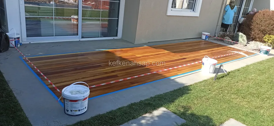 Newly laid deck on concrete in front of a glass door, edges masked with tape and a white paint bucket beside it