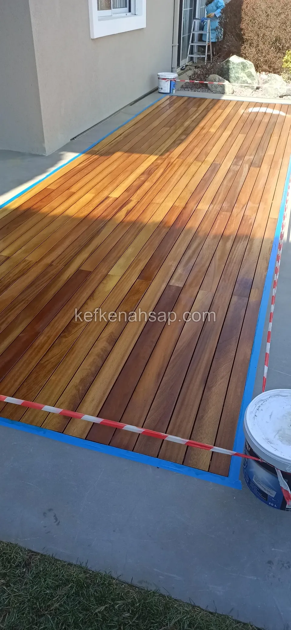 Ev yanındaki dar alanda mavi bantla kenarları korunmuş, yeni yağlanmış turuncu kahve deck ve önde boya kovası