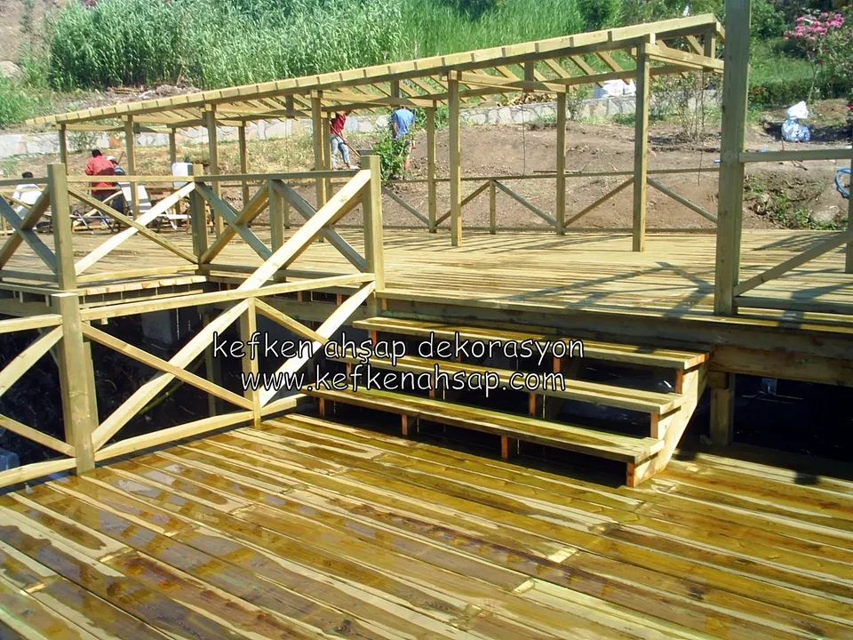 Eğimli arazide basamaklarla bağlanan iki seviyeli deck, çapraz X korkuluklar ve yükselen pergola kirişleri, çalışan ustalar
